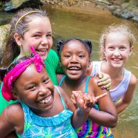 4 young girls hugging and smiling at the camera as they play in a creek.
