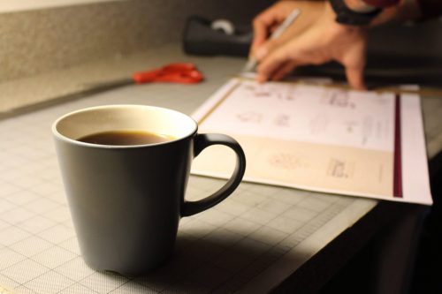 Close-up of designer's hands trimming a design on a cutting mat near a coffee cup.